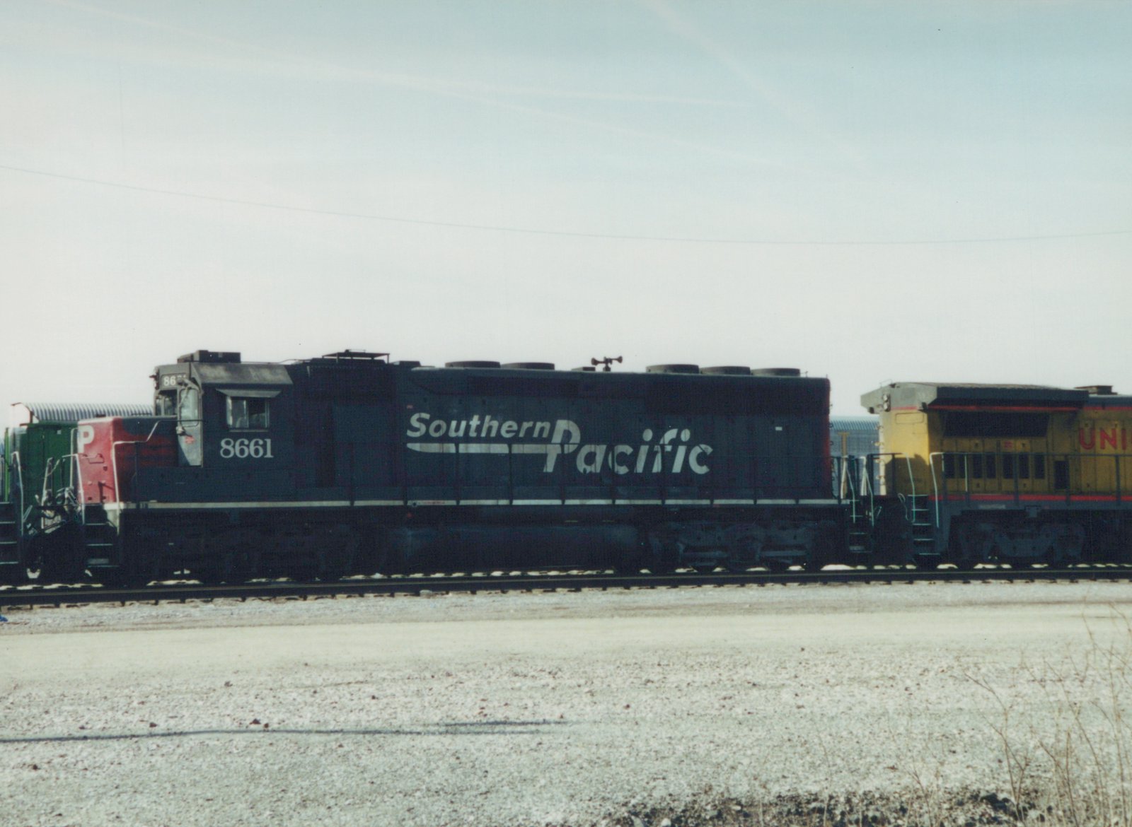 A side view of a black Southern Pacific diesel locomotive with the number 8661, parked in a gravel yard with other locomotives nearby under an overcast sky.