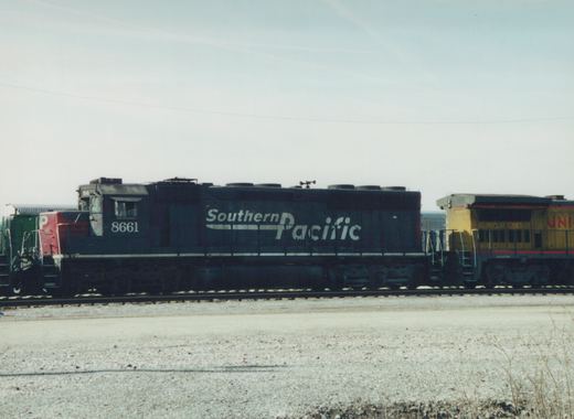 A side view of a black Southern Pacific diesel locomotive with the number 8661, parked in a gravel yard with other locomotives nearby under an overcast sky.