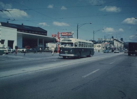 A vintage green and white city bus drives down a wide street lined with 1960s-era buildings and parked cars, with a large red 'Martin Wonderful Service' sign visible in the background.