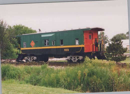 A side view of a green and black caboose with red cupola and yellow stripe, marked T.B.W. 523, parked on a railroad track surrounded by tall grass and trees.