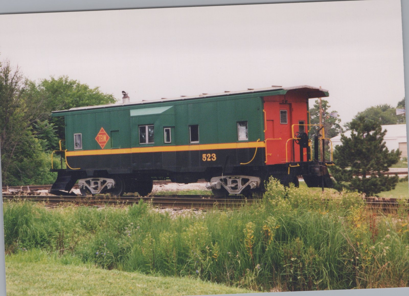A side view of a green and black caboose with red cupola and yellow stripe, marked T.B.W. 523, parked on a railroad track surrounded by tall grass and trees.