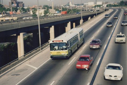 An articulated CTA bus drives on a multi-lane highway with several cars, under an elevated overpass on a sunny day.