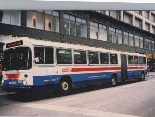 A white, red, and blue articulated bus with 'cta' lettering and the number 7109 on a city street with a building displaying 'METROPOLIS' and holiday lights in the background.