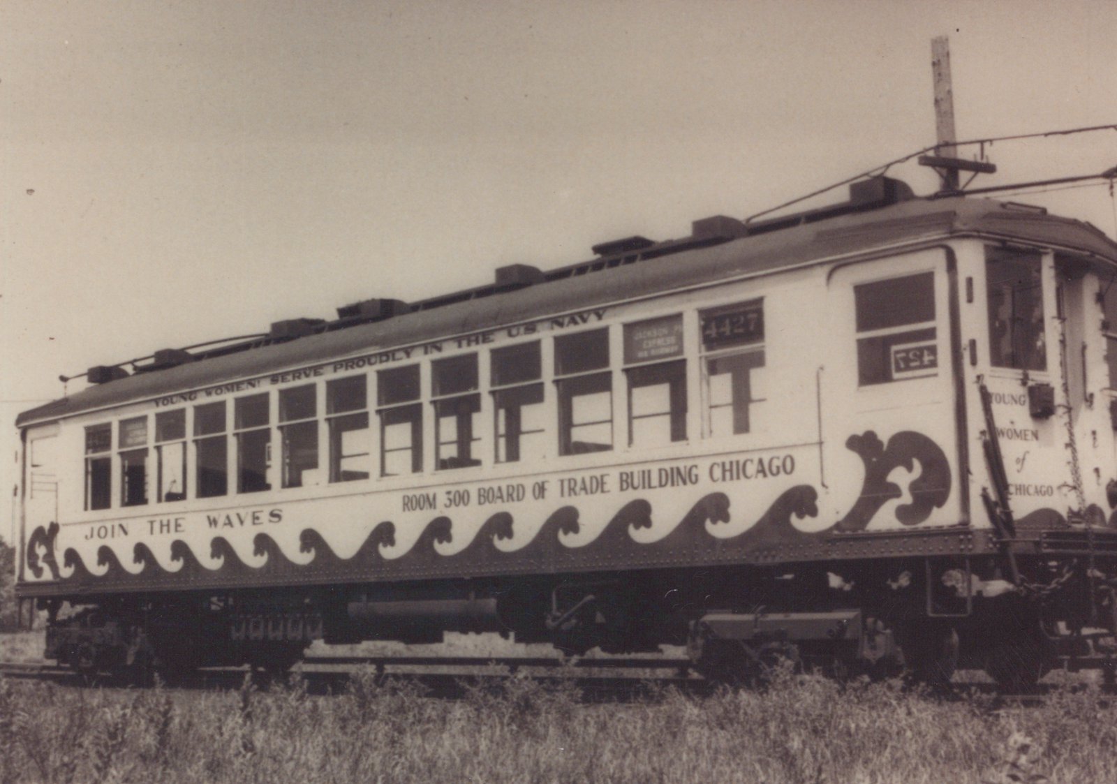 A vintage black and white photo shows a white, wood-bodied electric interurban car numbered 4427 with a navy recruitment ad along its side, sitting on tracks in a grassy area.