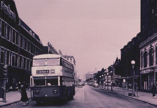 A vintage black-and-white photo shows a double-decker bus on a wide city street lined with multi-story buildings and period automobiles, with pedestrians walking on the sidewalks.