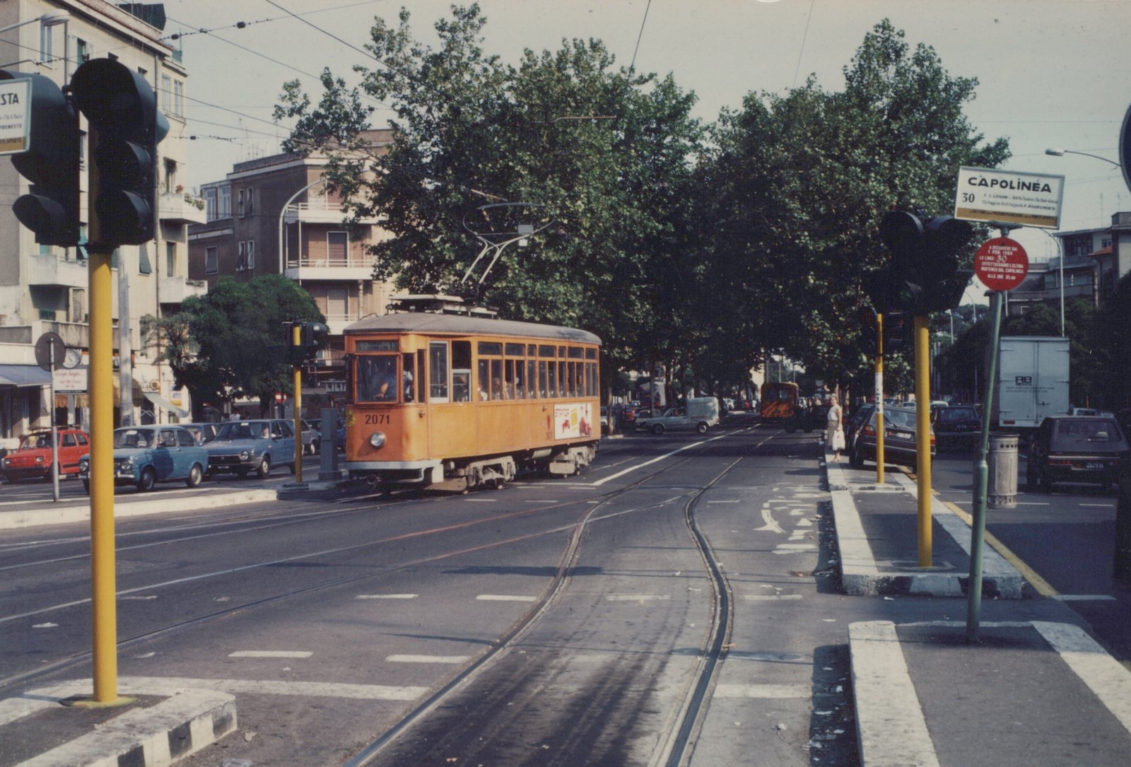 An orange streetcar on a sunlit city street in Rome, with a street sign reading 'CAPOLINEA 30' and a red sign announcing a service change effective in 1984.