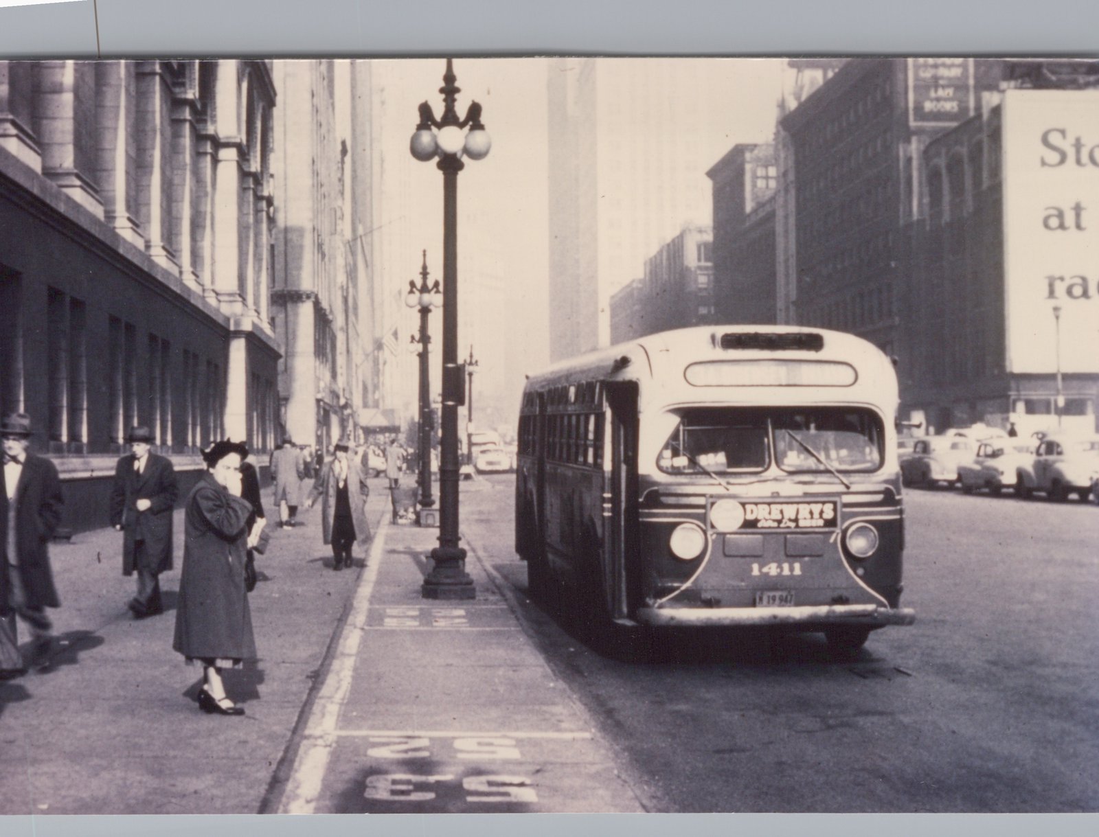 A black and white photograph of a vintage city bus with the number 1411 on its front, parked on a street with pedestrians walking on the sidewalk and tall buildings in the background.