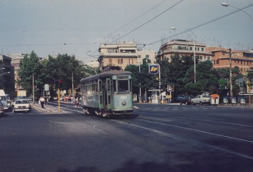 A vintage green streetcar with the number 2151 and route 30 sign is driving on a city street in Rome, passing a gas station and surrounded by cars and pedestrians on a sunny day.