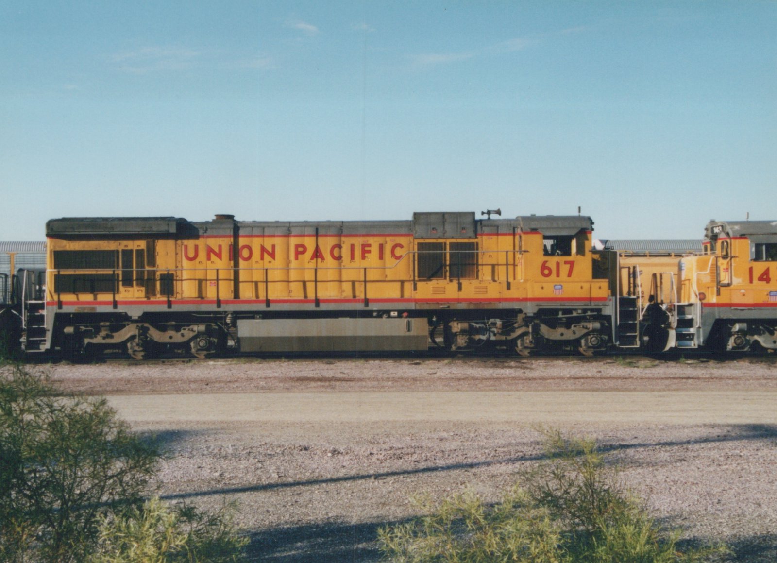 Side view of a yellow and gray Union Pacific locomotive with number 617 on its cab, standing in a gravel yard with another locomotive behind it, under a blue sky.
