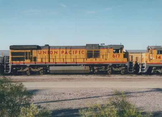 Side view of a yellow and gray Union Pacific locomotive with number 617 on its cab, standing in a gravel yard with another locomotive behind it, under a blue sky.