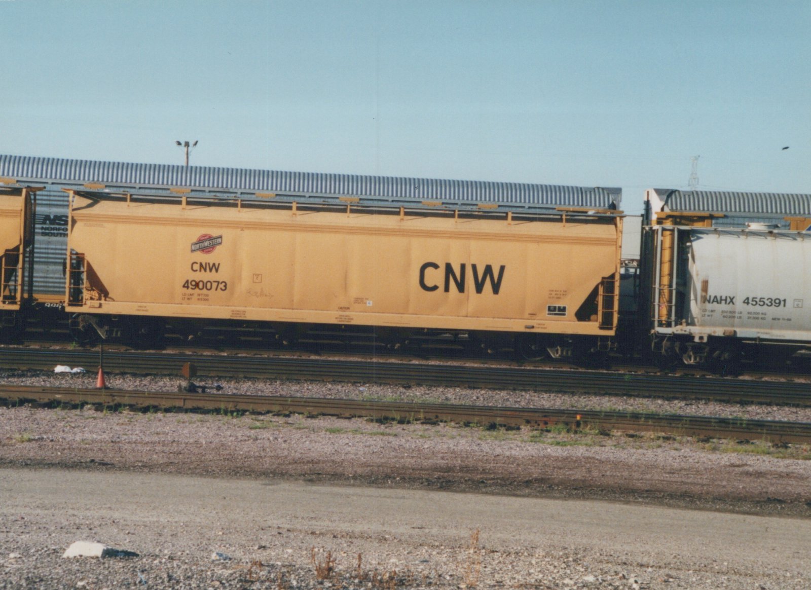 A yellow Chicago and North Western freight car numbered 490073 is shown in a rail yard alongside a white NAHX car and a dark Norfolk Southern car under a clear sky.