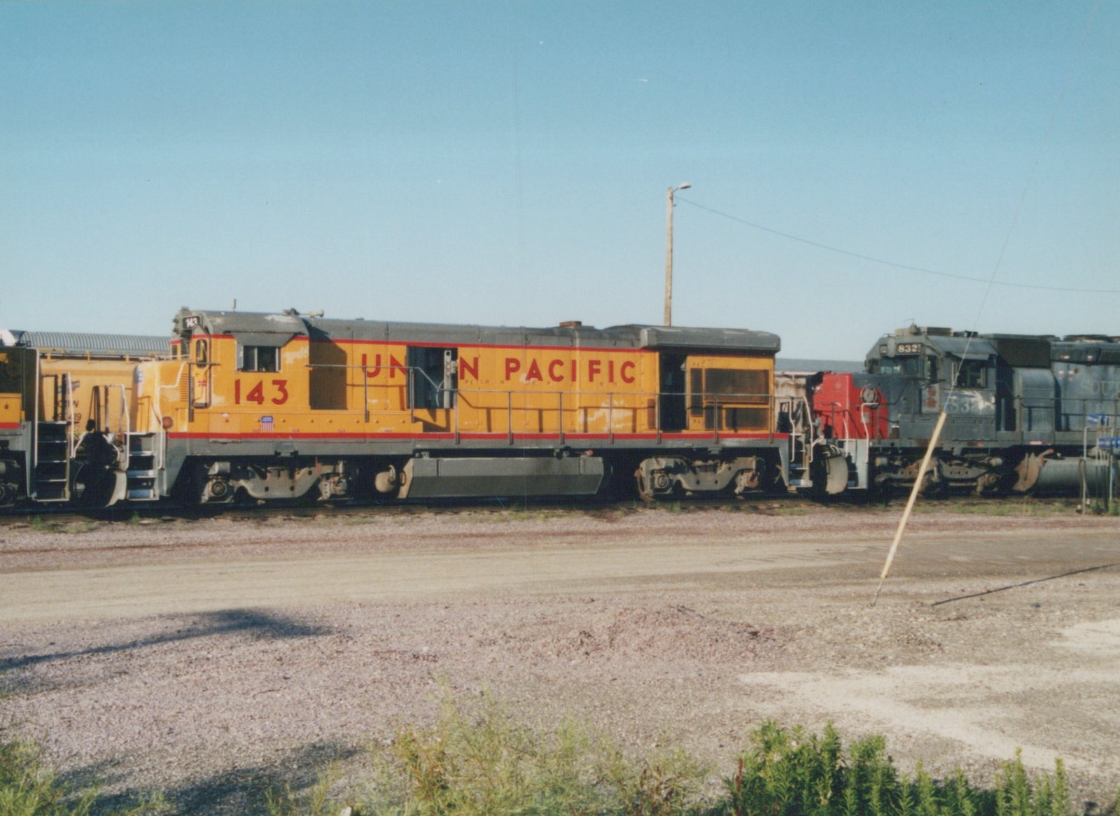 A yellow and red Union Pacific diesel locomotive numbered 143 is parked in a gravel yard, with another locomotive visible in the background.