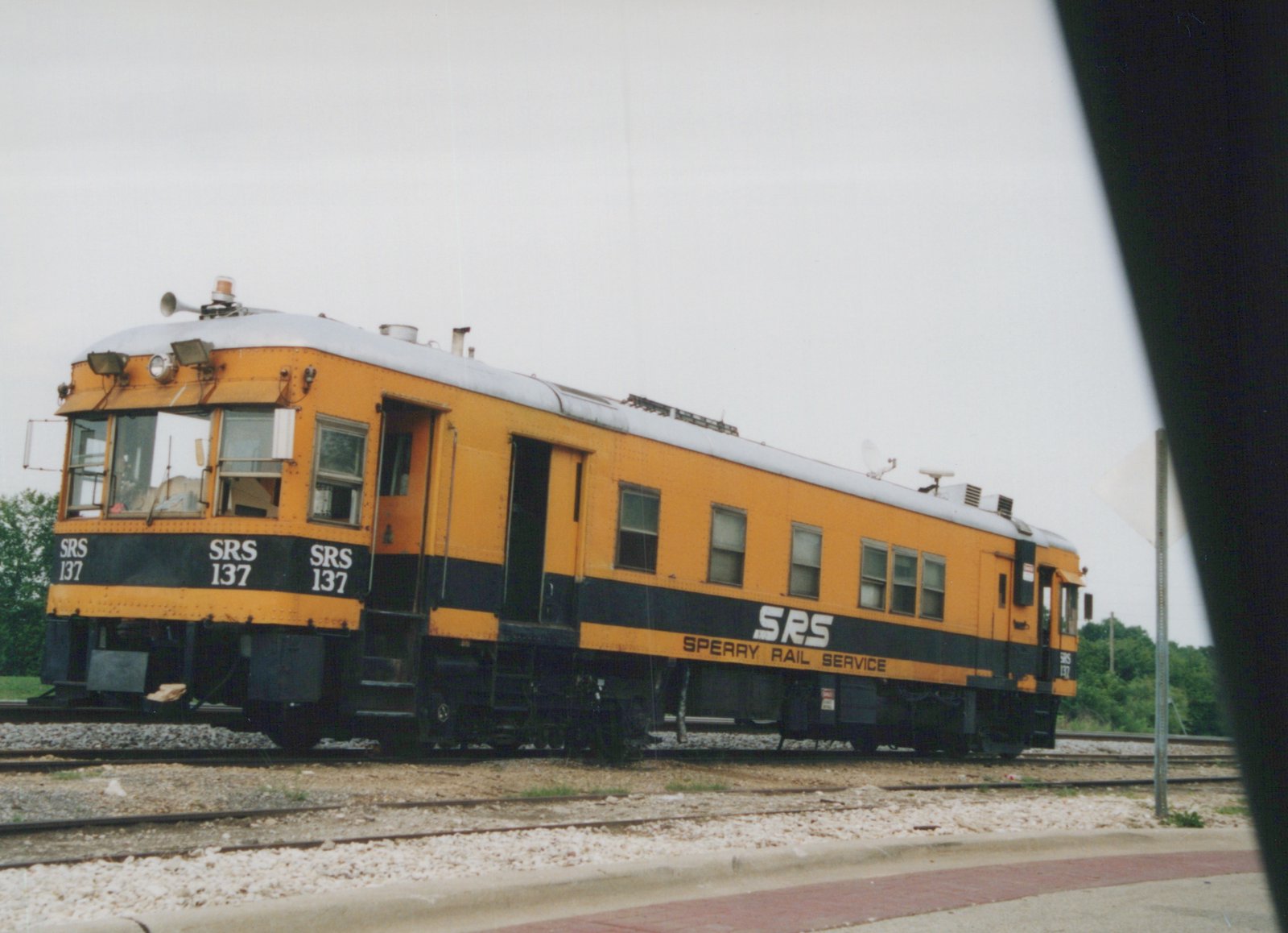 A yellow and black self-propelled railcar with 'SRS 137' and 'Sperry Rail Service' lettering is parked on railroad tracks under an overcast sky.