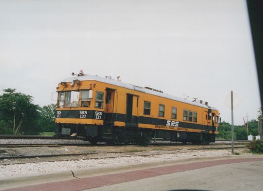 A yellow and black self-propelled railcar with 'SRS 137' on the front and 'Sperry Rail Service' on the side, sitting on tracks next to a paved road with a red brick sidewalk, under a cloudy sky.