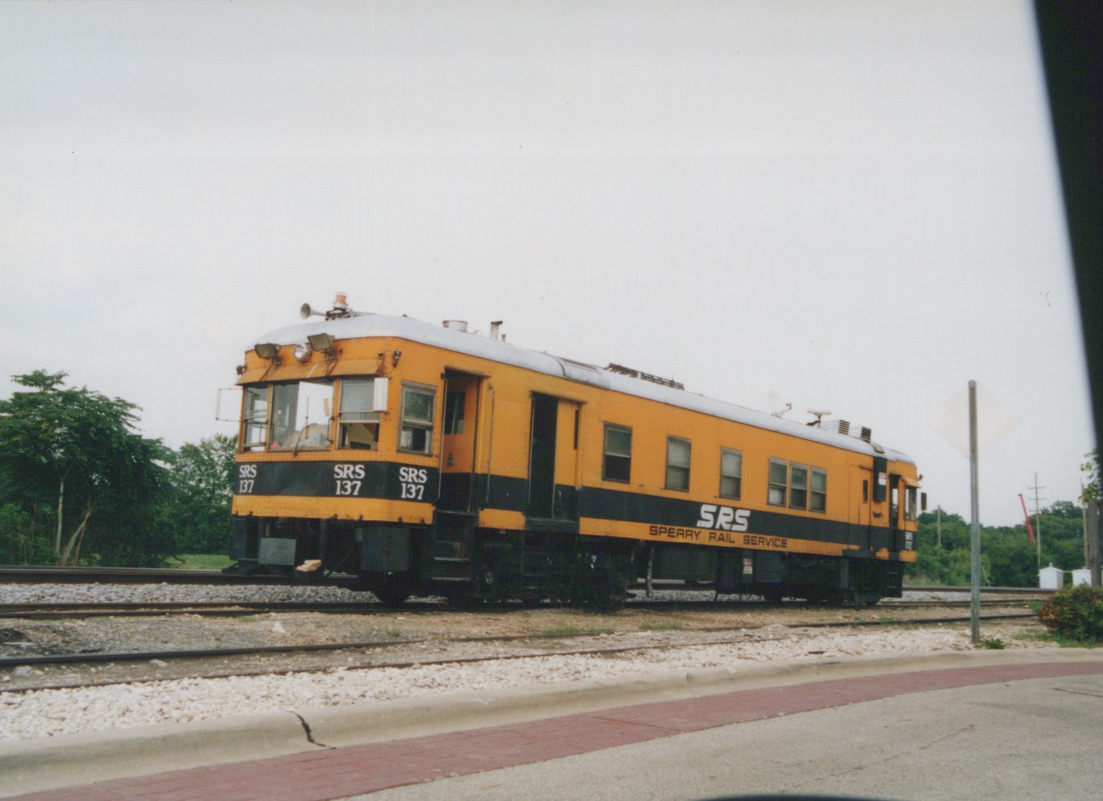 A yellow and black self-propelled railcar with 'SRS 137' on the front and 'Sperry Rail Service' on the side, sitting on tracks next to a paved road with a red brick sidewalk, under a cloudy sky.