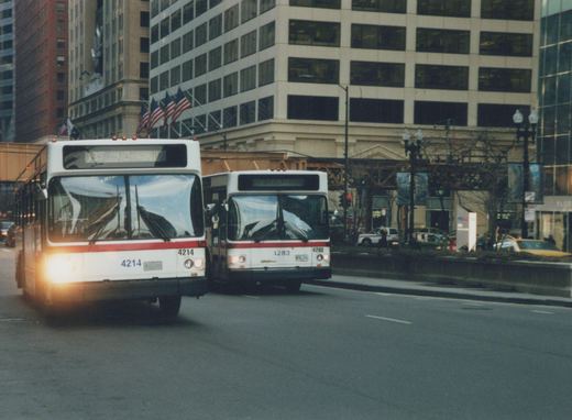 Two white and red CTA buses, numbers 4214 and 4283, drive on a city street with tall buildings and a few pedestrians in the background.