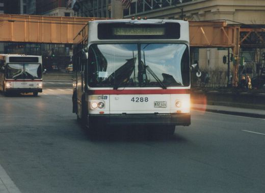 Front view of a white and red Chicago Transit Authority bus, number 4288, with its headlights on, driving on a city street under an elevated train track, with another CTA bus visible in the background.
