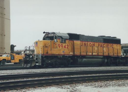 A yellow and red Union Pacific locomotive numbered 9942 is parked on multiple tracks in a rail yard under a gray sky.