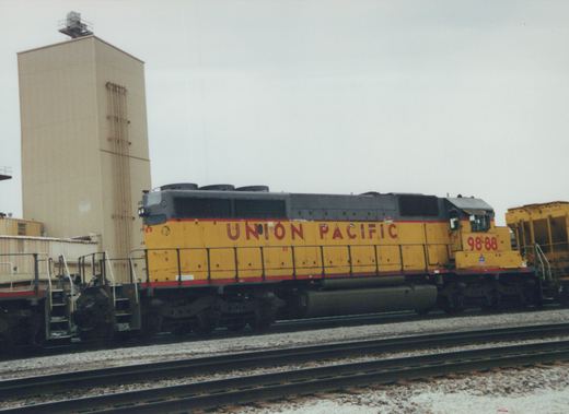 A side view of a yellow and gray Union Pacific locomotive numbered 9888 parked on tracks in a rail yard with a tall beige building in the background.