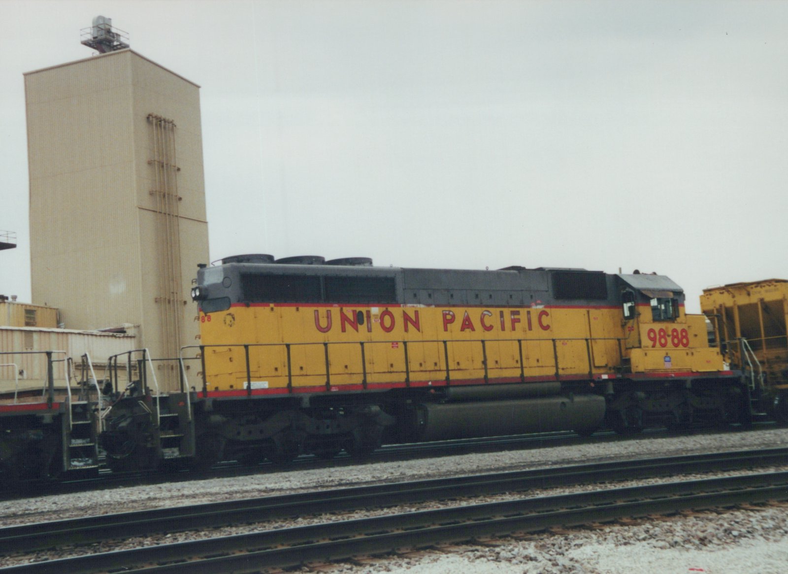 A side view of a yellow and gray Union Pacific locomotive numbered 9888 parked on tracks in a rail yard with a tall beige building in the background.