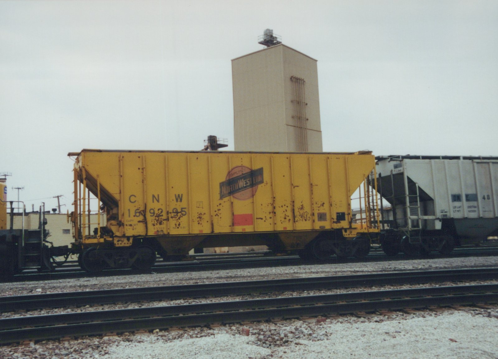 Side view of a yellow Chicago and North Western freight car on multiple tracks in a yard, with a grain silo visible behind it and a white Burlington Northern car next to it.