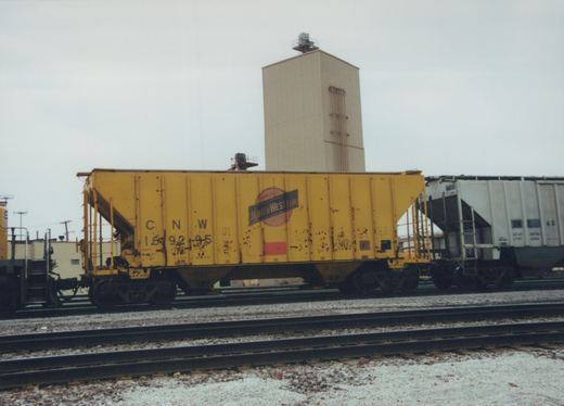 Side view of a yellow Chicago and North Western freight car on multiple tracks in a yard, with a grain silo visible behind it and a white Burlington Northern car next to it.