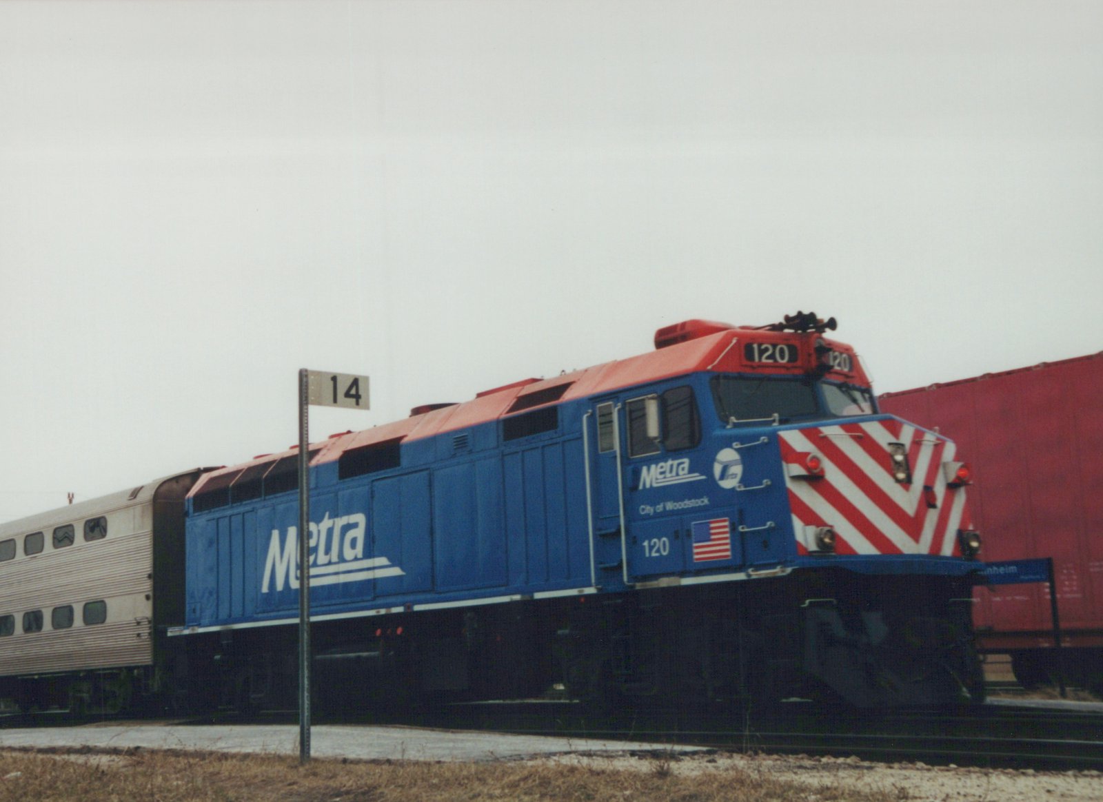 A blue and red Metra diesel locomotive numbered 120, with "City of Woodstock" lettering, is shown pulling a silver commuter car on a cloudy, snowy day.