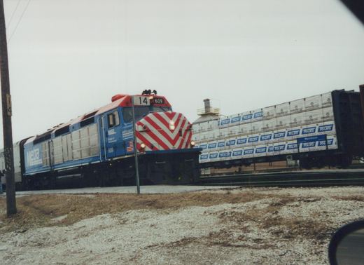 A blue and red diesel locomotive with the number 609 and Metra lettering is pulling a train of freight cars loaded with lumber past a snowy roadside.