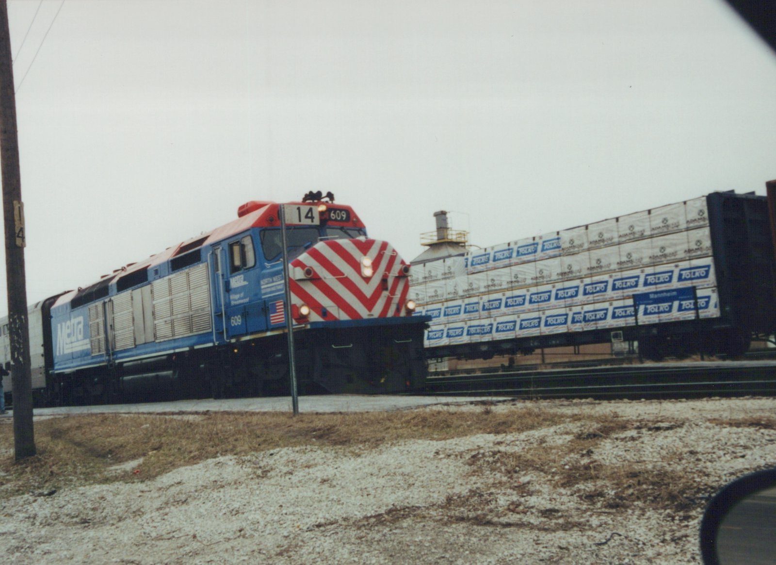 A blue and red diesel locomotive with the number 609 and Metra lettering is pulling a train of freight cars loaded with lumber past a snowy roadside.