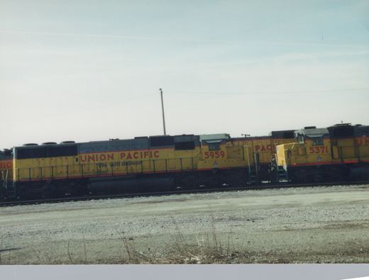 Four yellow and gray Union Pacific diesel locomotives, numbered 5959 and 5371 among others, are parked side by side in a gravel yard under an overcast sky.