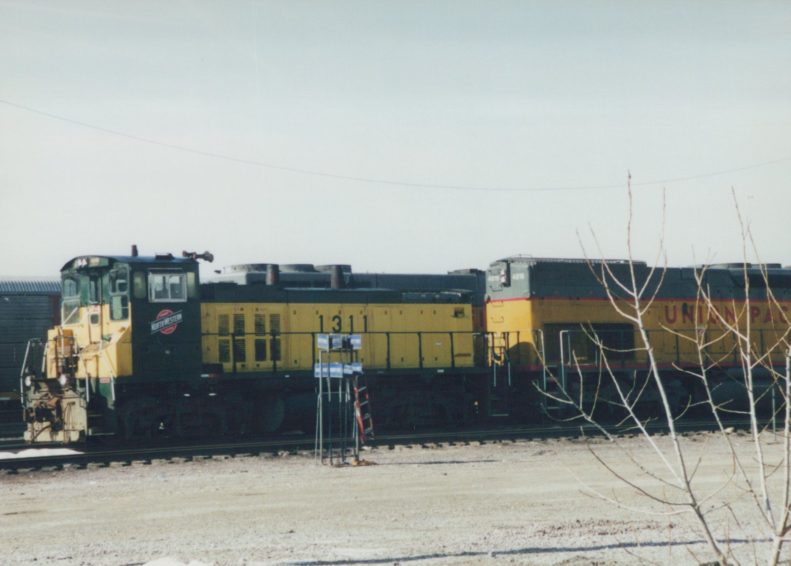 Two diesel locomotives, one from Chicago and North Western and one from Union Pacific, are parked side-by-side in a gravel yard with bare trees in the foreground.