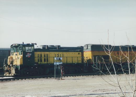 Two diesel locomotives, one from Chicago and North Western and one from Union Pacific, are parked side-by-side in a gravel yard with bare trees in the foreground.