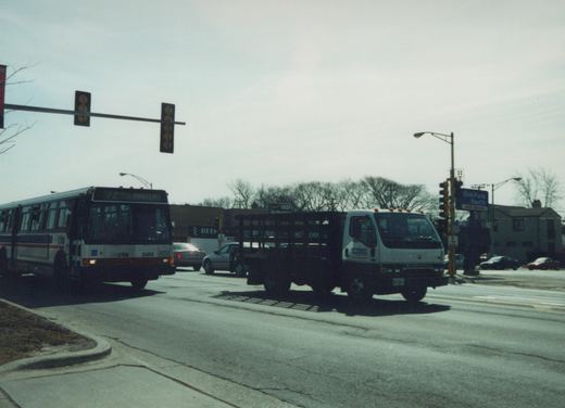 A white Chicago Transit Authority bus with red and blue stripes, numbered 5469, is turning at an intersection in Oak Park with a white flatbed truck and several cars nearby, under a cloudy sky with bare trees.