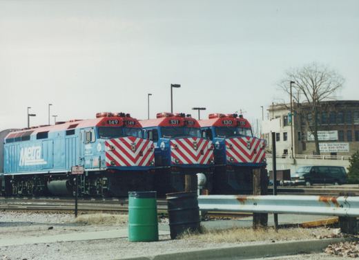 Three blue and red Metra locomotives are parked next to each other in a yard, with a building and a minivan in the background.