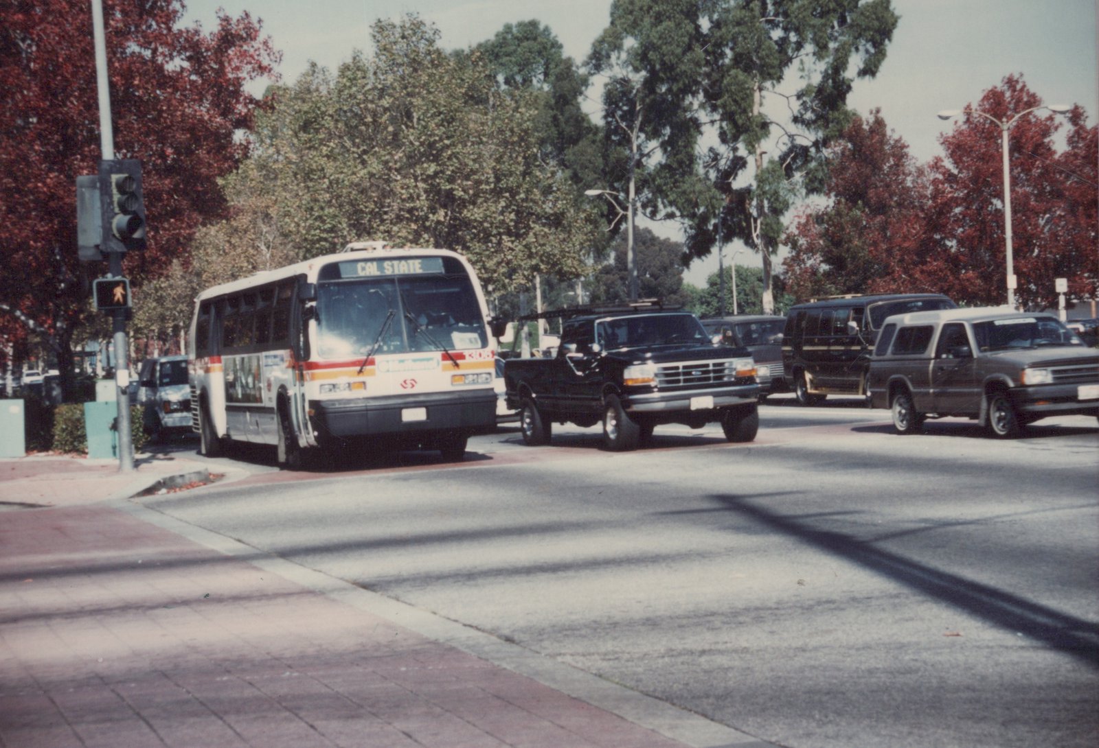 A white Southwestern bus with red and orange stripes, numbered K105, is stopped at a street intersection with its 'CAL STATE' destination sign displayed, alongside cars from the 1990s, under clear skies with autumn trees in the background.