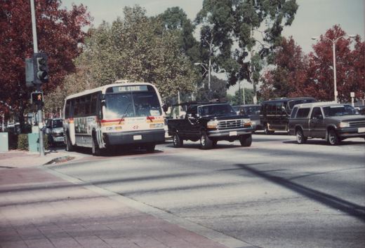 A white Southwestern bus with red and orange stripes, numbered K105, is stopped at a street intersection with its 'CAL STATE' destination sign displayed, alongside cars from the 1990s, under clear skies with autumn trees in the background.