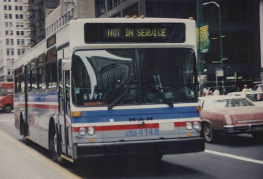 A white, blue, and red Chicago Transit Authority bus, number 4148, with 'NOT IN SERVICE' displayed on its front sign, is driving on a city street with other cars and tall buildings nearby.