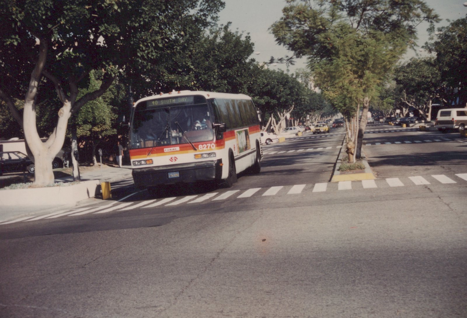 A white and orange GMC bus with the number 8270 is driving down a city street lined with trees and parked cars on a sunny day.