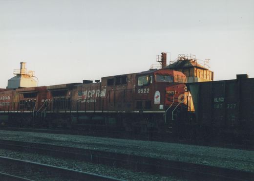 Side view of two dark brown CP Rail System diesel locomotives with red cabs, numbered 9522 and 9671, coupled together on railroad tracks next to a green freight car marked HLMX, with industrial buildings in the background under a clear evening sky.