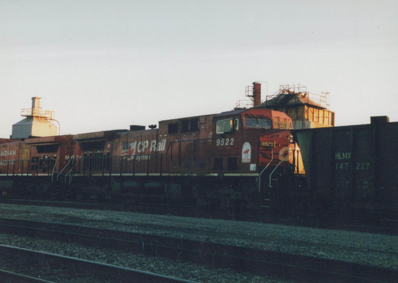 Side view of two dark brown CP Rail System diesel locomotives with red cabs, numbered 9522 and 9671, coupled together on railroad tracks next to a green freight car marked HLMX, with industrial buildings in the background under a clear evening sky.