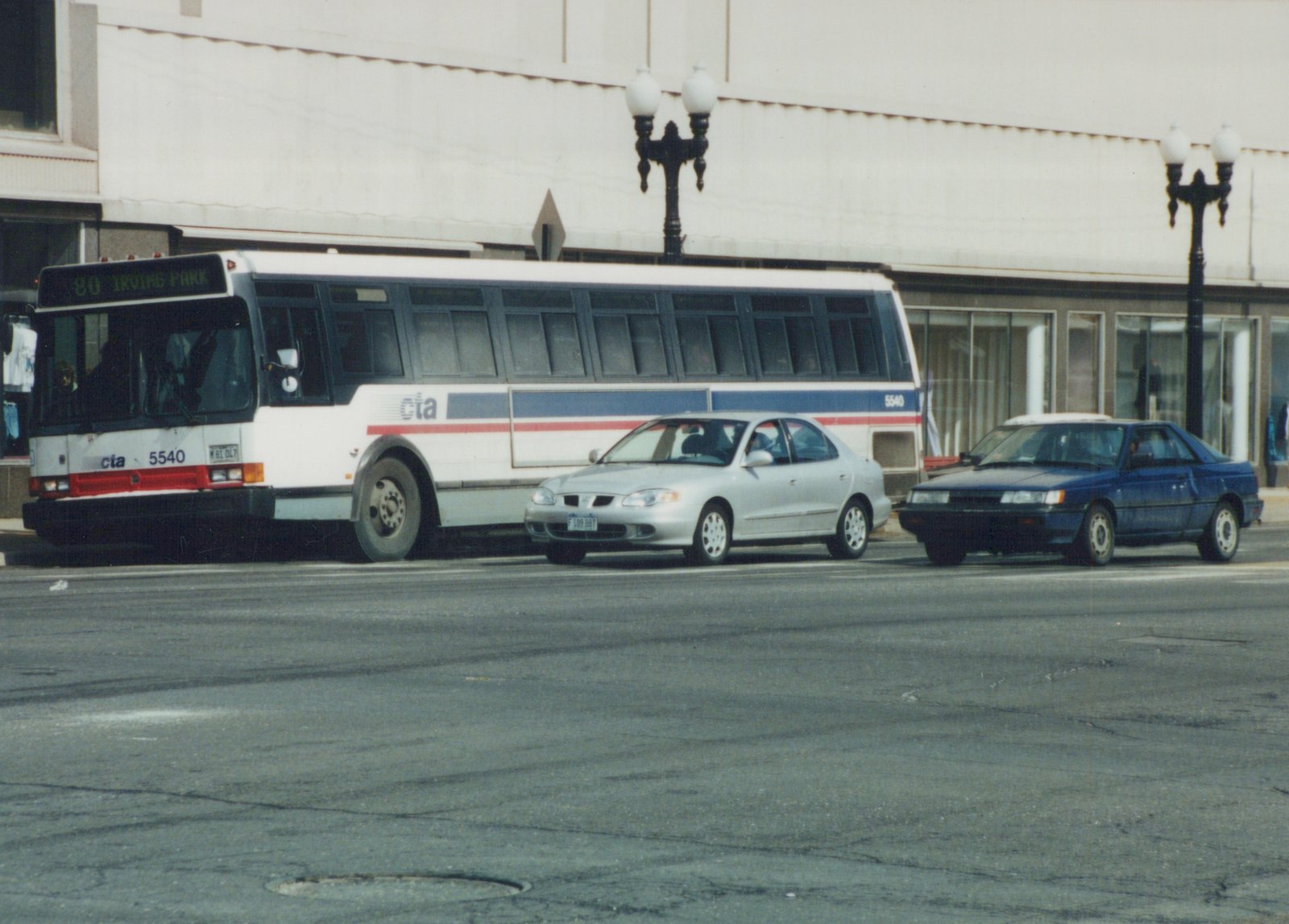 A white, blue and red Chicago Transit Authority bus, number 5540, on route 80 to Irving Park, is parked on a street in Cicero, Illinois, with a silver car and a blue car beside it.