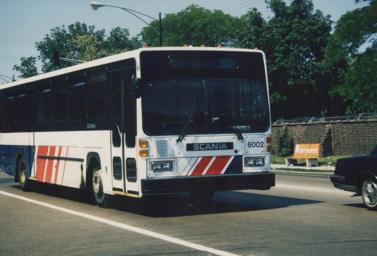 A white, blue, and red Chicago Transit Authority bus, number 6002, is driving down a city street on a sunny day with green trees and a brick wall in the background.