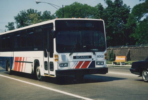 A white, blue, and red Chicago Transit Authority bus, number 6002, is driving down a city street on a sunny day with green trees and a brick wall in the background.