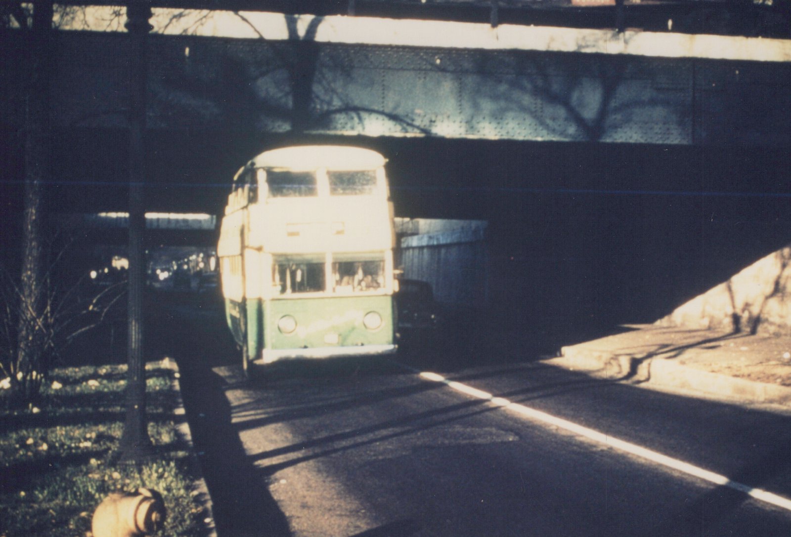 Front view of a vintage green and cream double-decker-style bus under a dark steel railroad bridge, with bare trees and a clear sky.
