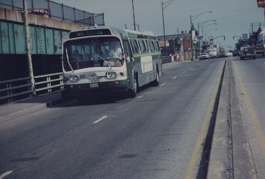 A green and cream CTA bus, fleet number 7453, driving on a multi-lane road next to a bridge structure with other cars and pedestrians visible in the background.