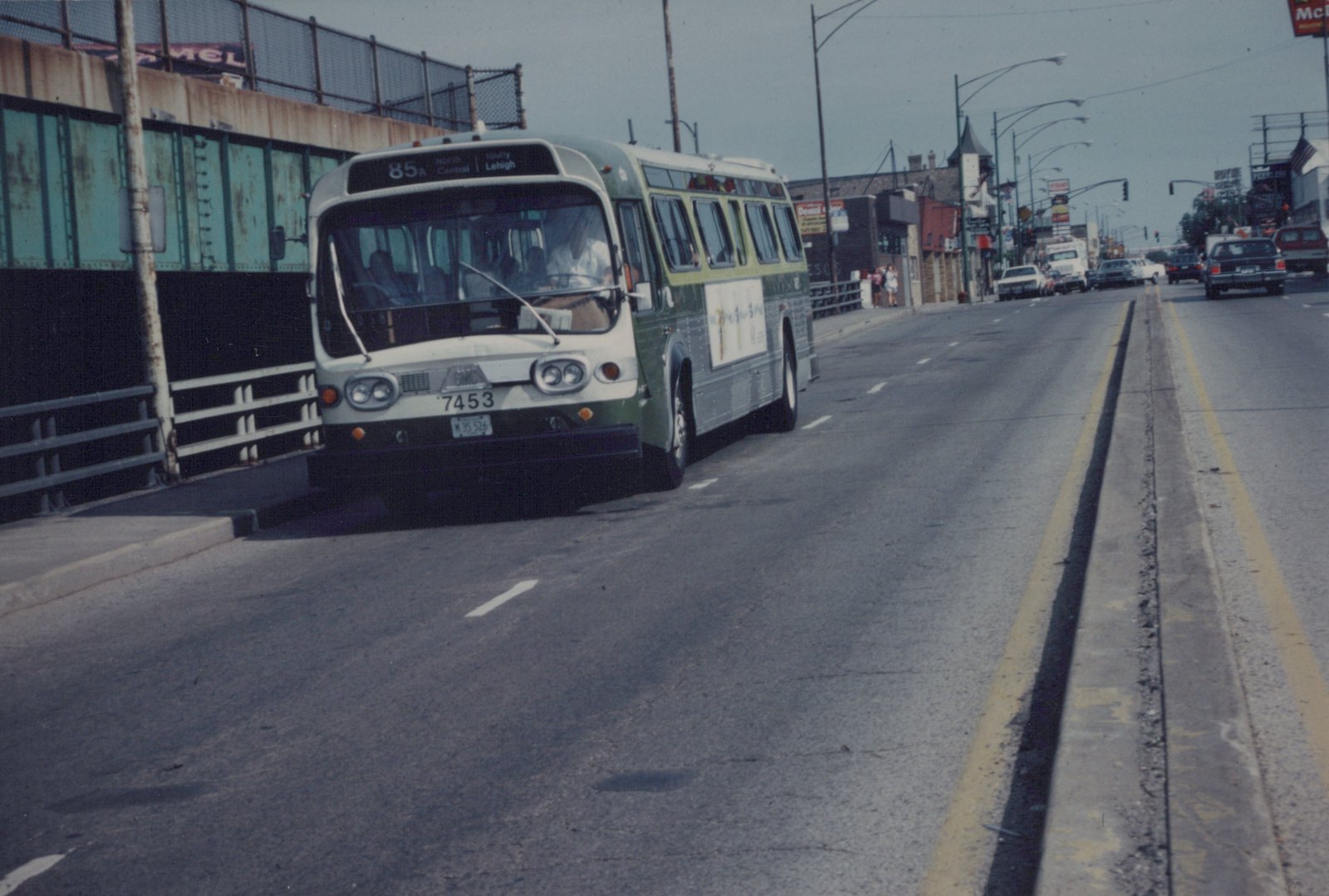 A green and cream CTA bus, fleet number 7453, driving on a multi-lane road next to a bridge structure with other cars and pedestrians visible in the background.