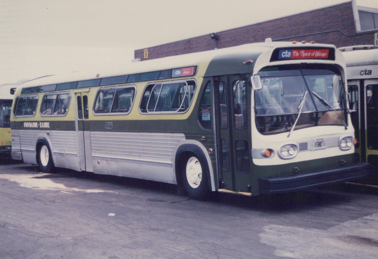 A green, cream, and silver CTA bus with the number P999 parked in a lot, with other buses and a brick building behind it.
