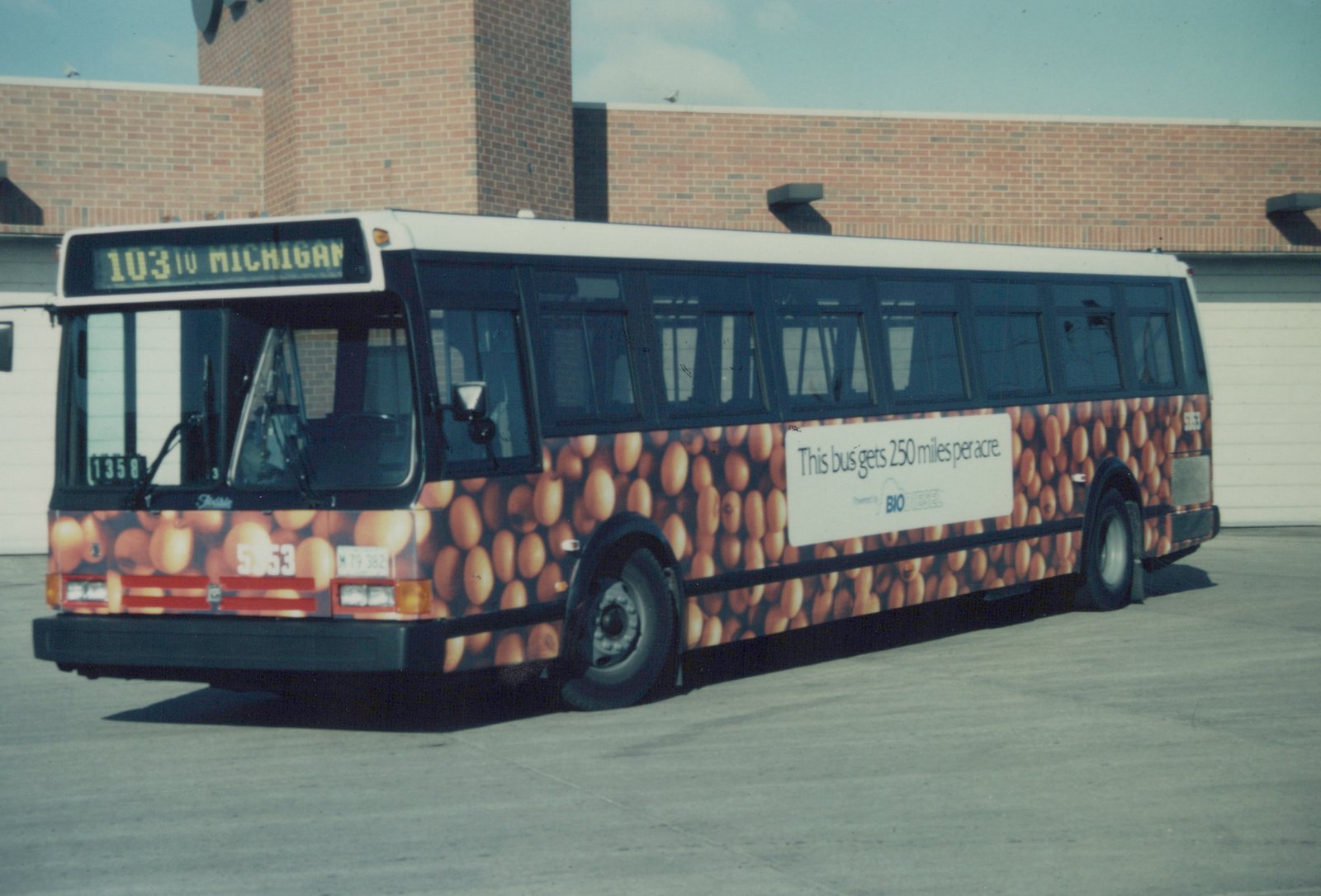 A white-roofed city bus with a soybean-themed advertisement is parked on a paved lot in front of a brick building, showing its destination sign and fleet number.