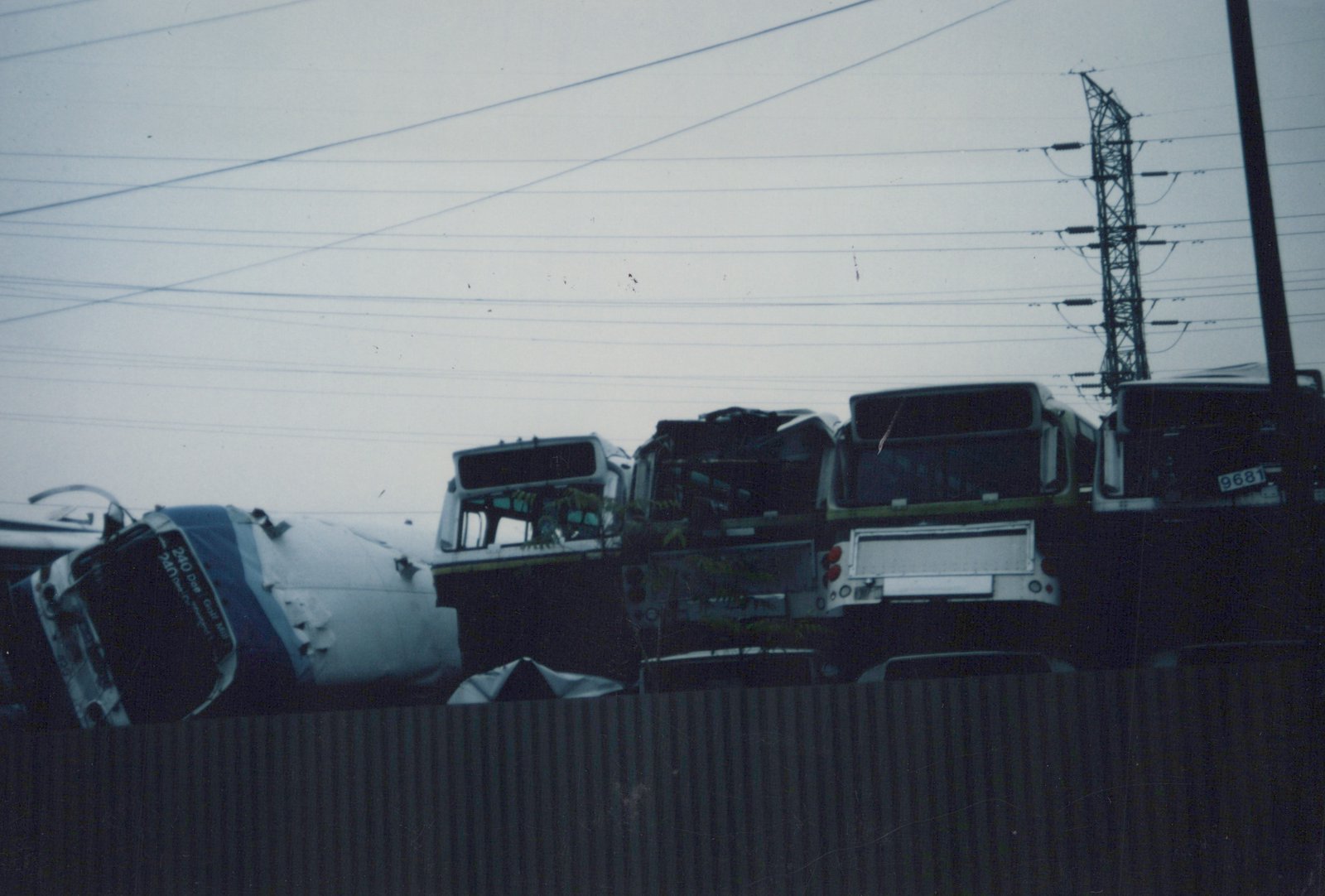 A stack of junked buses, mostly in dark green and white livery, in a salvage yard with overhead power lines and a steel tower visible against a gray sky.