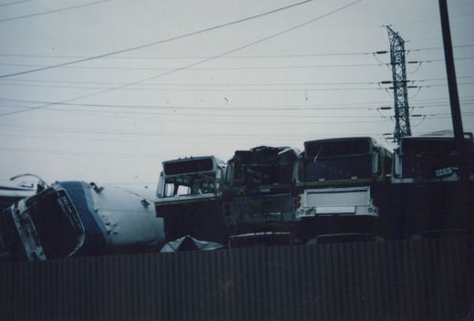 A stack of junked buses, mostly in dark green and white livery, in a salvage yard with overhead power lines and a steel tower visible against a gray sky.